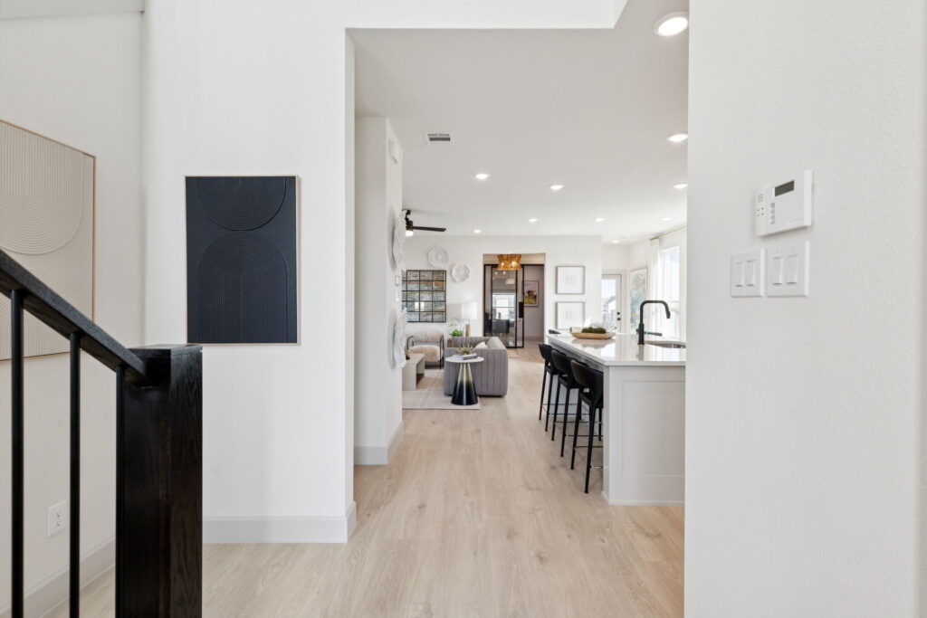 Modern open-concept living space in Painted Tree featuring light wood flooring, white walls, a kitchen island with barstools, and a view of the living room and windows in the background—showcasing the elegance of Normandy Homes’ new homes.