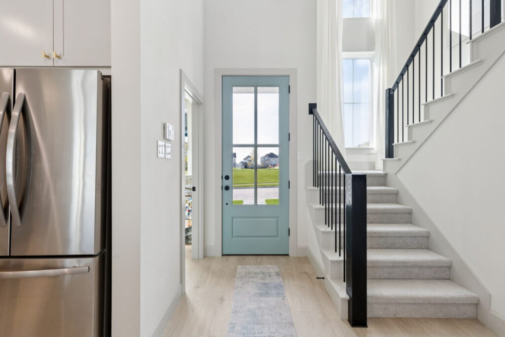 Entryway with a light blue front door in a Normandy Homes residence, adjacent stairs with black railing, white walls, and a stainless steel refrigerator on the left—showcasing the charm of new homes in Painted Tree.