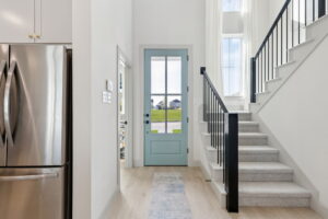 Entryway with a light blue front door in a Normandy Homes residence, adjacent stairs with black railing, white walls, and a stainless steel refrigerator on the left—showcasing the charm of new homes in Painted Tree.