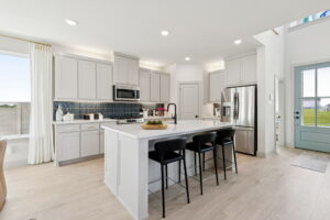 Modern kitchen in Normandy Homes' new homes at Painted Tree features white cabinets, a center island with three black stools, stainless steel appliances, and light wood flooring. Bright natural light streams through large windows and a glass door.