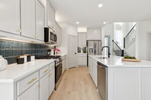 Modern kitchen in a McKinney new home by Normandy Homes, featuring white cabinets, stainless steel appliances, black tile backsplash, an island with sink, light wood flooring, plus stairs and front door visible in the background.