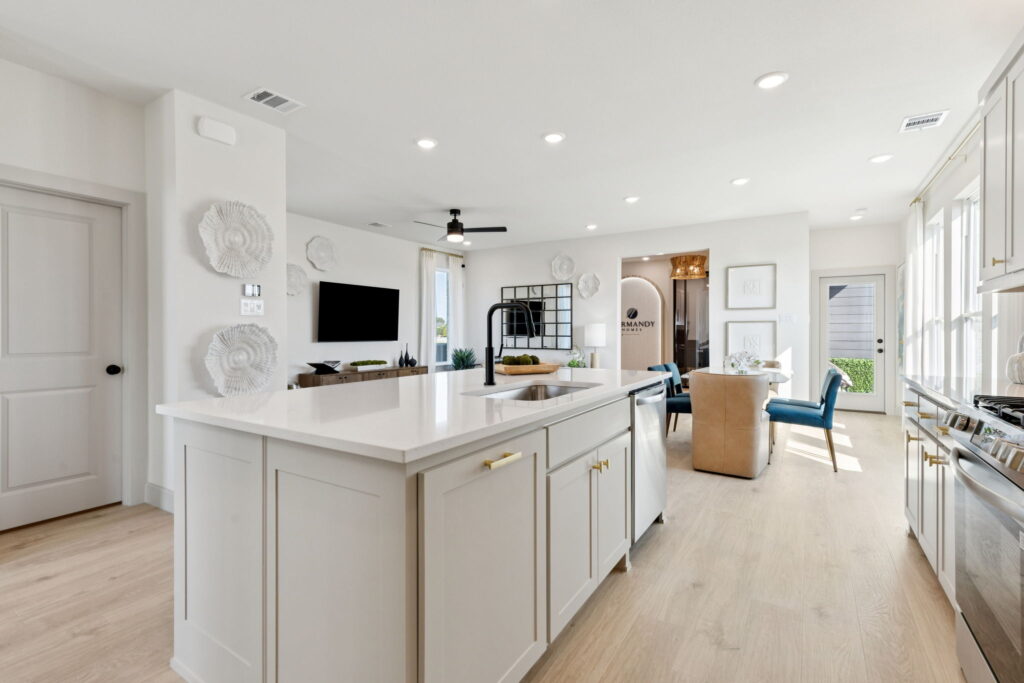 Bright, modern kitchen by Normandy Homes in McKinney’s Painted Tree, featuring a large white island, gold hardware, open shelving, and adjacent living and dining areas filled with natural light from large windows.