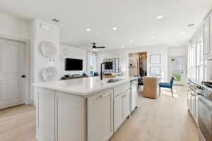 Bright, modern kitchen by Normandy Homes in McKinney’s Painted Tree, featuring a large white island, gold hardware, open shelving, and adjacent living and dining areas filled with natural light from large windows.