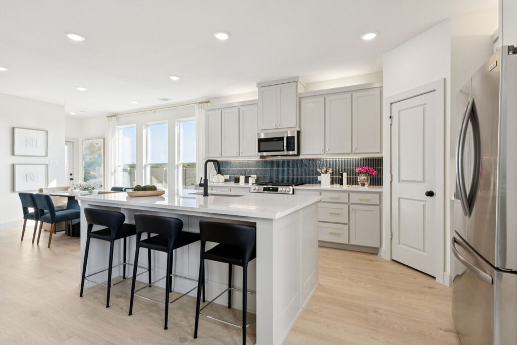 Modern kitchen with white cabinets, stainless steel appliances, a central island with black bar stools, and a dining area—experience stylish living in Normandy Homes’ new homes at Painted Tree.