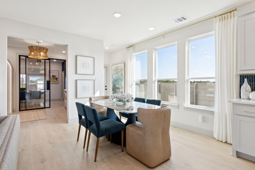Modern dining area in Painted Tree with a marble table, blue velvet and tan leather chairs, large windows, light wood flooring, and a view into the hallway with glass double doors—perfect for stylish new homes by Normandy Homes.
