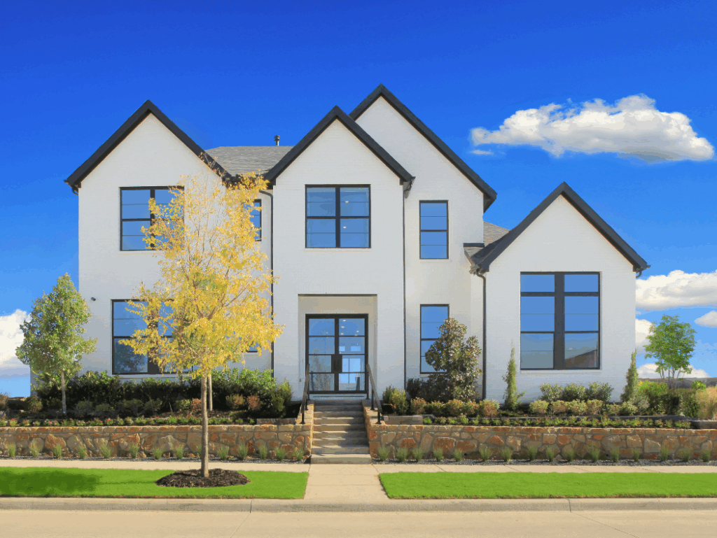 Modern two-story white house by Drees Custom Homes with large black-framed windows, landscaped front yard, and stone retaining wall, nestled in Painted Tree, TX, under a clear blue sky with a few clouds.