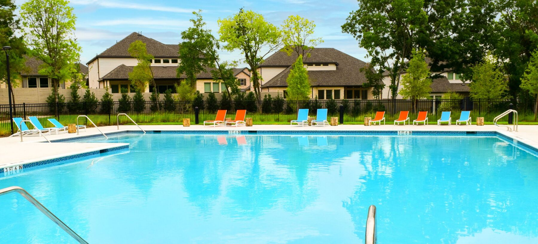 Outdoor swimming pool with clear blue water, surrounded by colorful lounge chairs, metal handrails, and houses and trees in the background under a blue sky—perfect for relaxing afternoons in McKinney TX. Located near Painted Tree’s Lakeside Trailhead, this inviting spot blends comfort with scenic community charm.